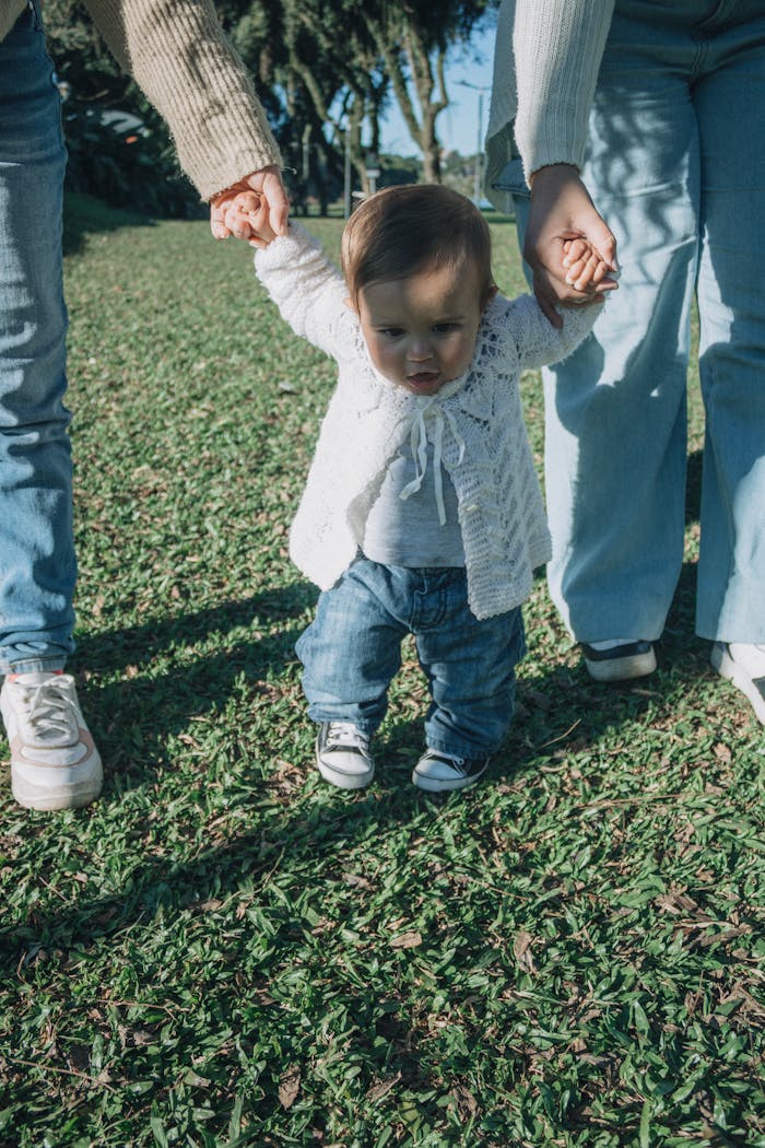 services-03 A baby takes first steps on grass, supported by two adults on a sunny day.